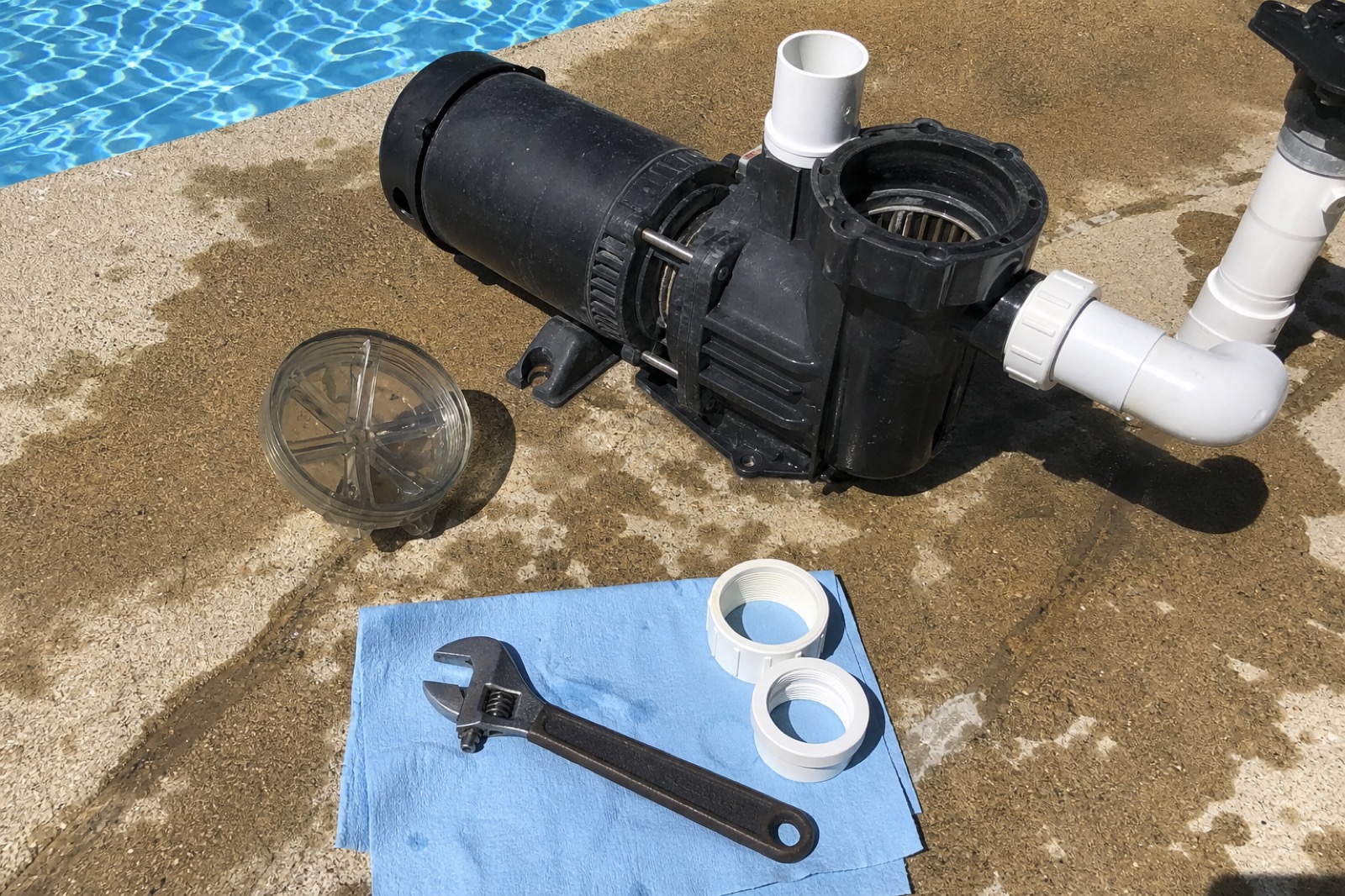 Disassembled pool pump on a concrete deck with adjustable wrench, strainer lid, and PVC couplings on a blue shop towel.