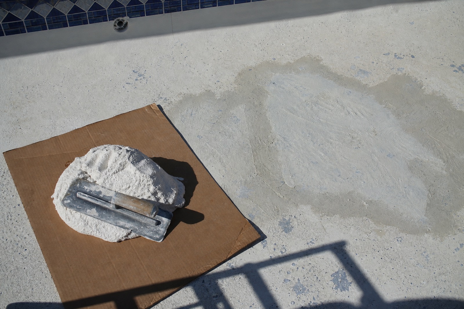 Fresh white pool plaster on cardboard with a trowel resting on top, beside an older plaster patch being worked on at the bottom of an empty pool.