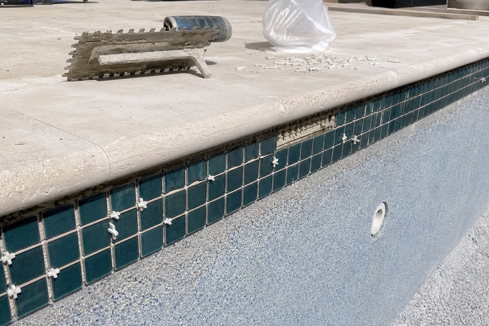 A band of dark teal mosaic tile being installed at the waterline of a new Sparkle Pool Care pool, with thinset trowel and spacers nearby.